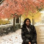 Priya Ravi wearing a winter coat, standing outside in the light snow near a tree with fall colored leaves.